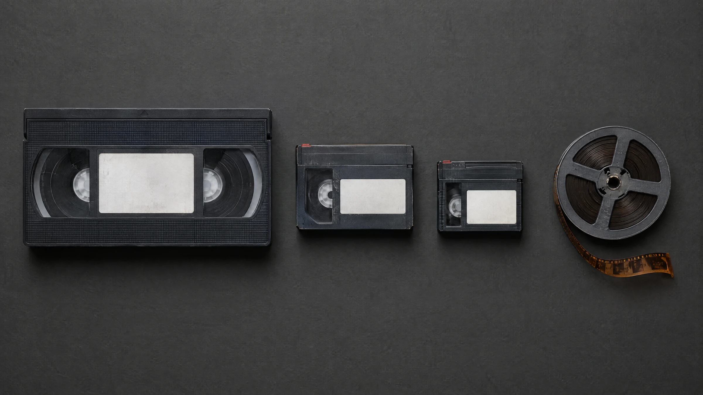 Illustration: a full-size VHS cassette, two smaller camcorder cassettes and a small film reel with a strip of film unspooled, laid out on a dark grey surface