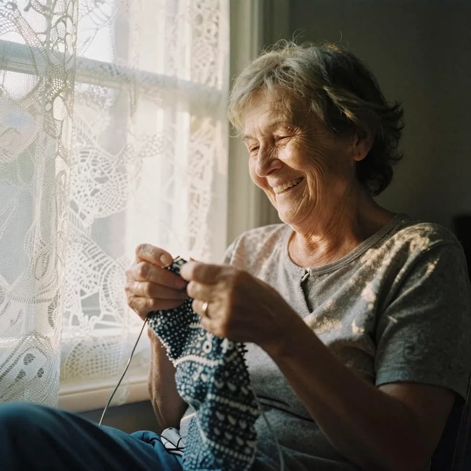 An older woman knitting by a lace-curtained window, deep laugh lines and paper-thin skin lit by warm afternoon sun filtering through the fab