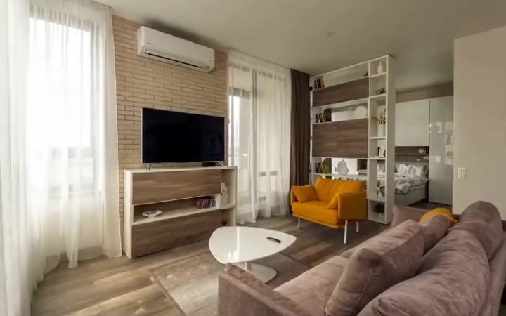 A frame from a reel generated from that same listing photo: the identical living space — pale brick wall, wall-mounted TV, mustard-yellow armchair, grey sofa and the bed nook behind the open shelving — with the camera now inside the room