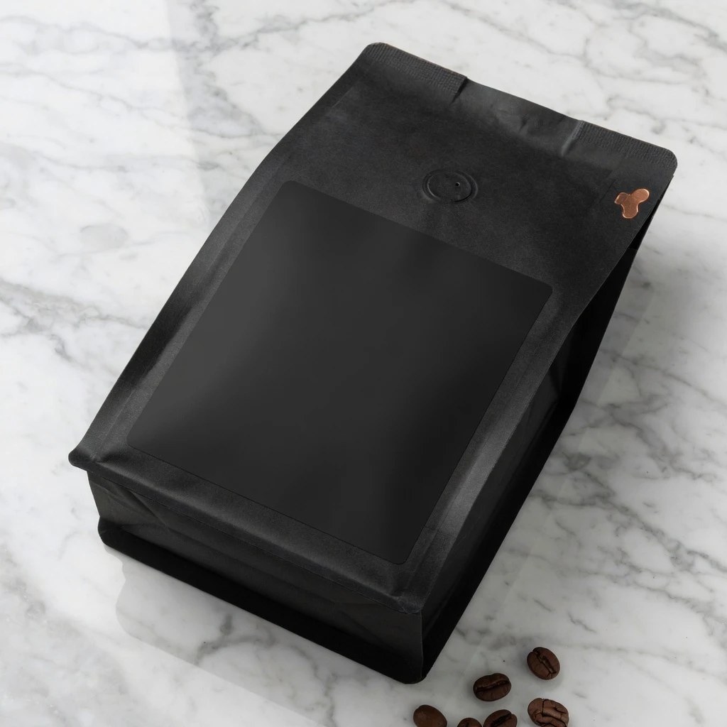 A matte black coffee pouch on a marble counter with a blank, unprinted label panel on the front and a few beans beside it