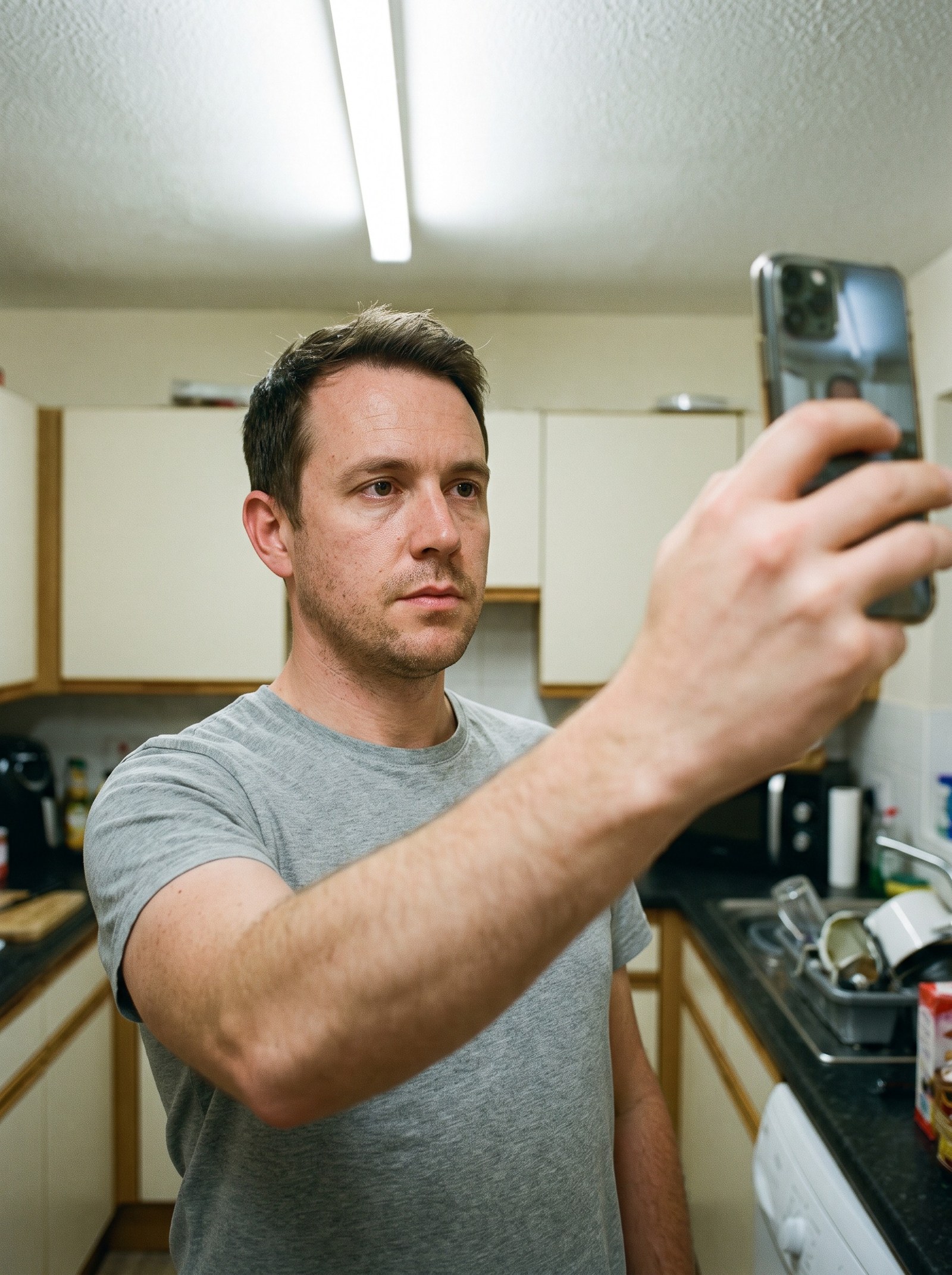 A casual phone selfie of a man in a plain grey t-shirt, taken at arm's length in a kitchen under flat overhead light