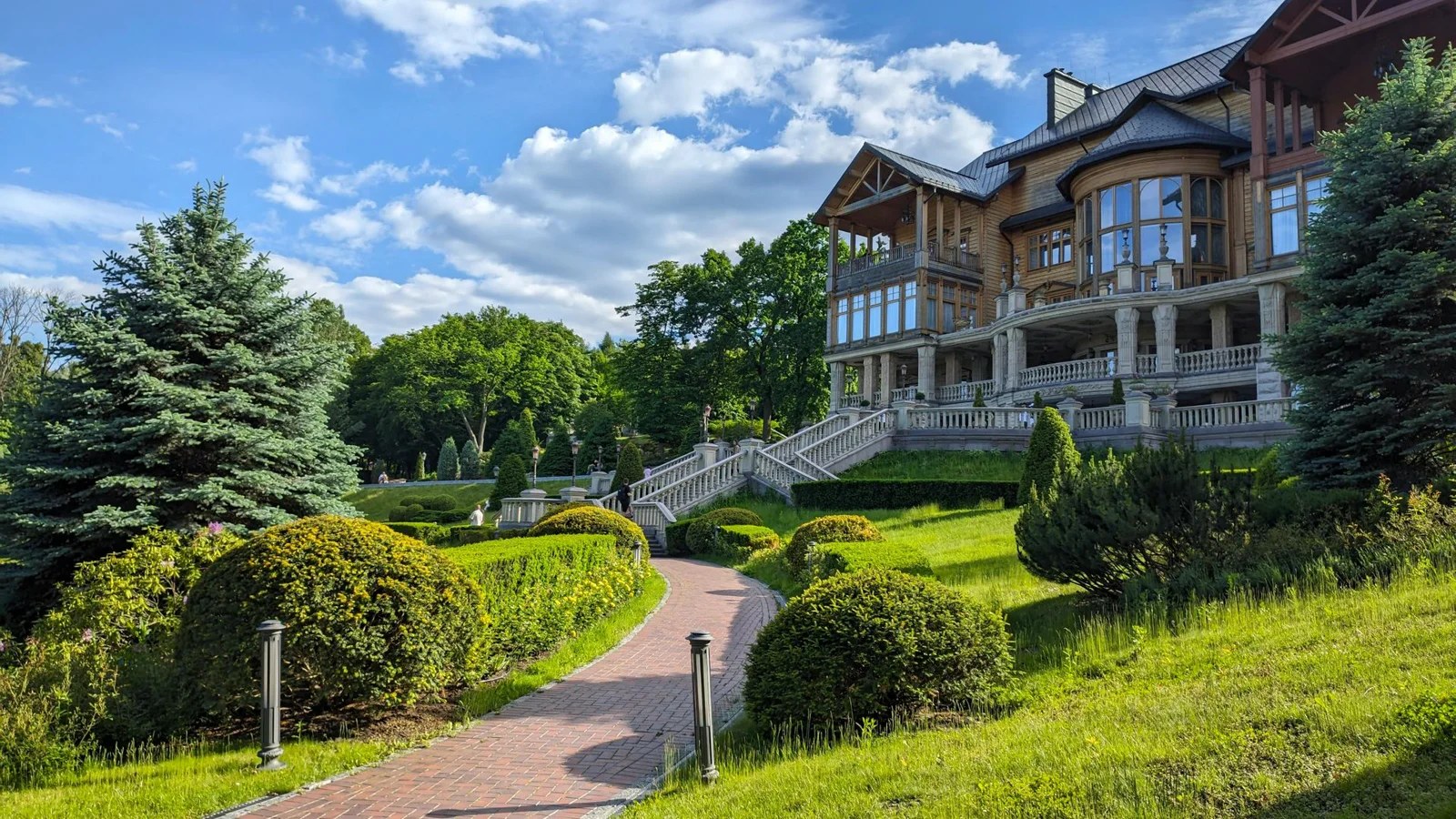 The source photograph: a timber lodge with a glazed corner tower standing above a clipped formal garden, a brick path curving toward its stone staircase