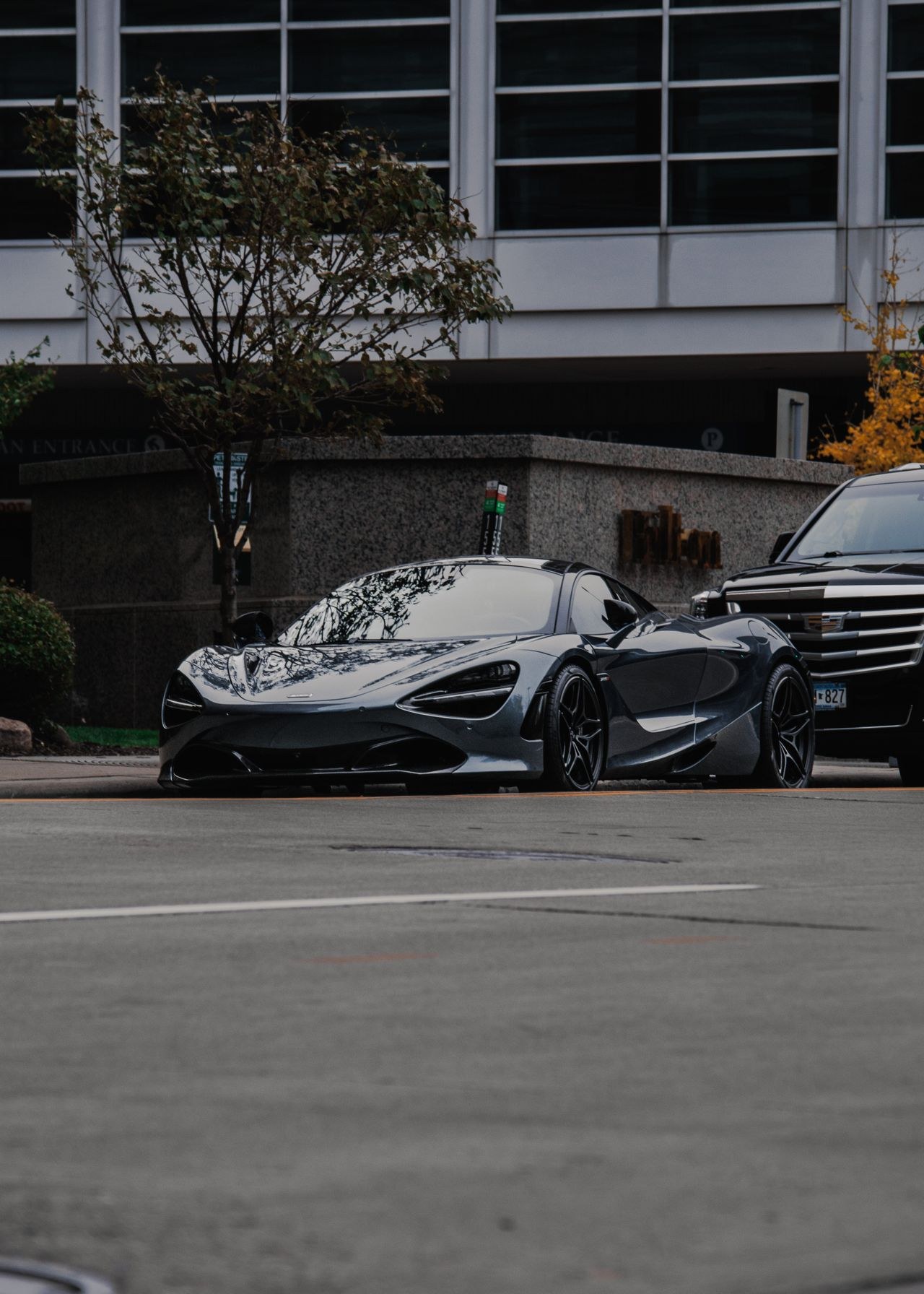 The source photograph as it was taken: a low, wide grey supercar at the kerb outside a city building in flat overcast daylight, a black SUV parked alongside it