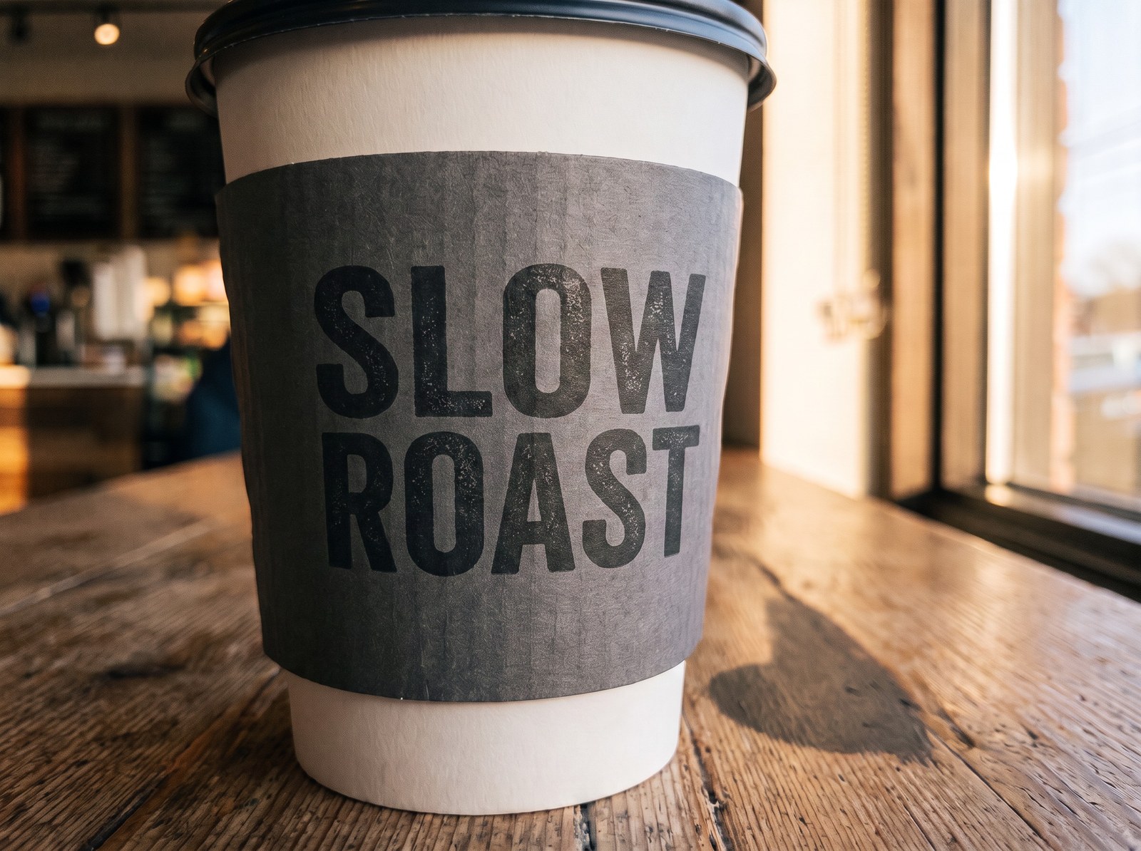 The same cup, sleeve, table and window light after the edit: the sleeve now reads SLOW ROAST in the same heavy condensed uppercase face, the same charcoal ink and the same slightly rough printed-on-card finish, set as the same two-line block