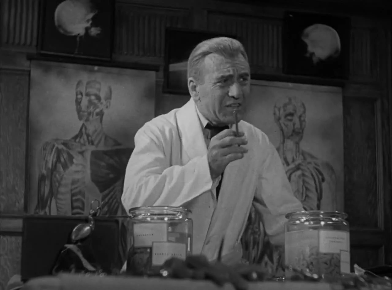 A frame from the source clip: a black-and-white scene of a grey-haired man in a lab coat pausing with a pipe in hand, anatomical charts and specimen jars behind him, speaking his original language