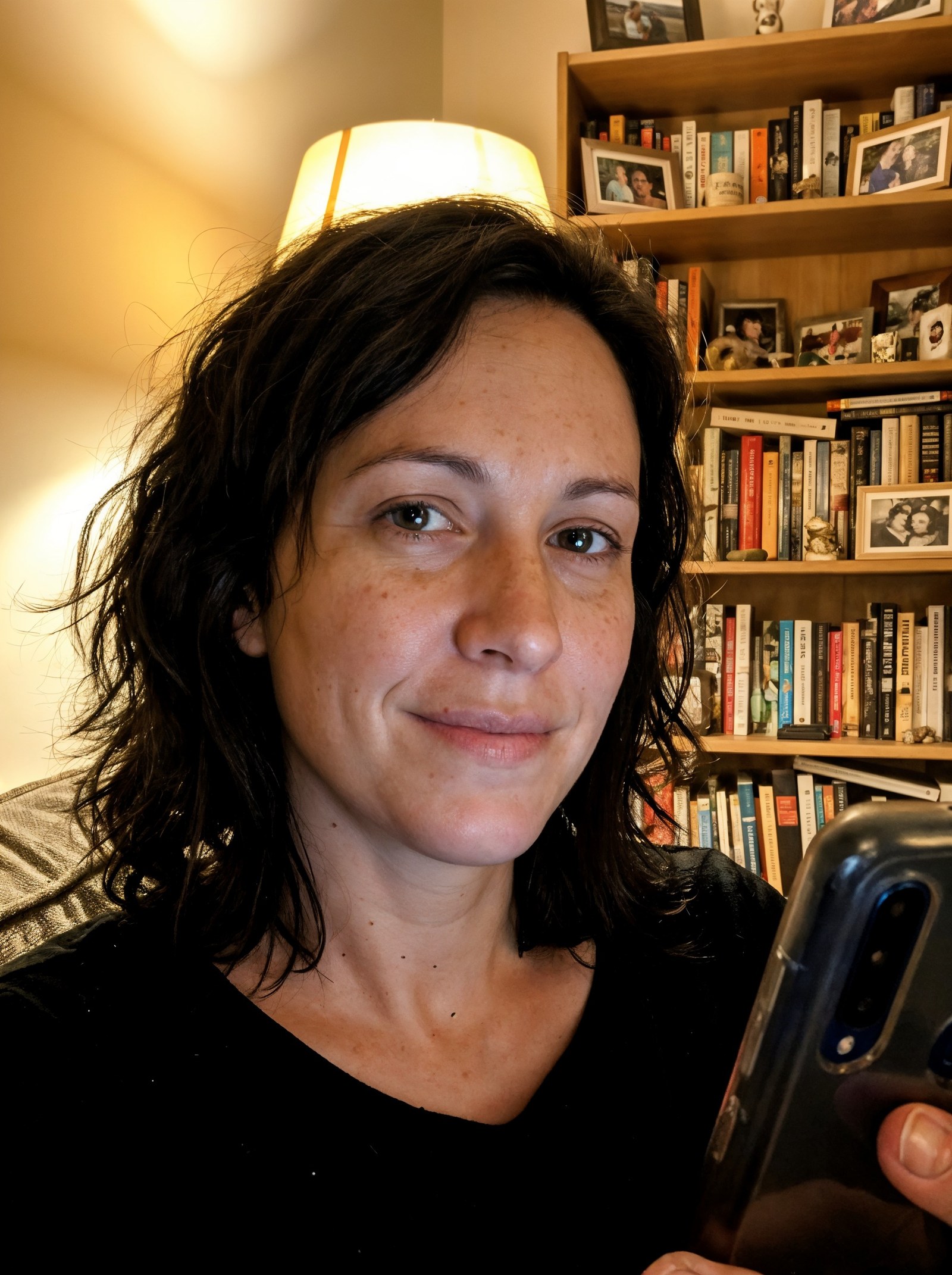 An off-centre phone selfie taken in warm lamp light, a cluttered bookshelf behind her