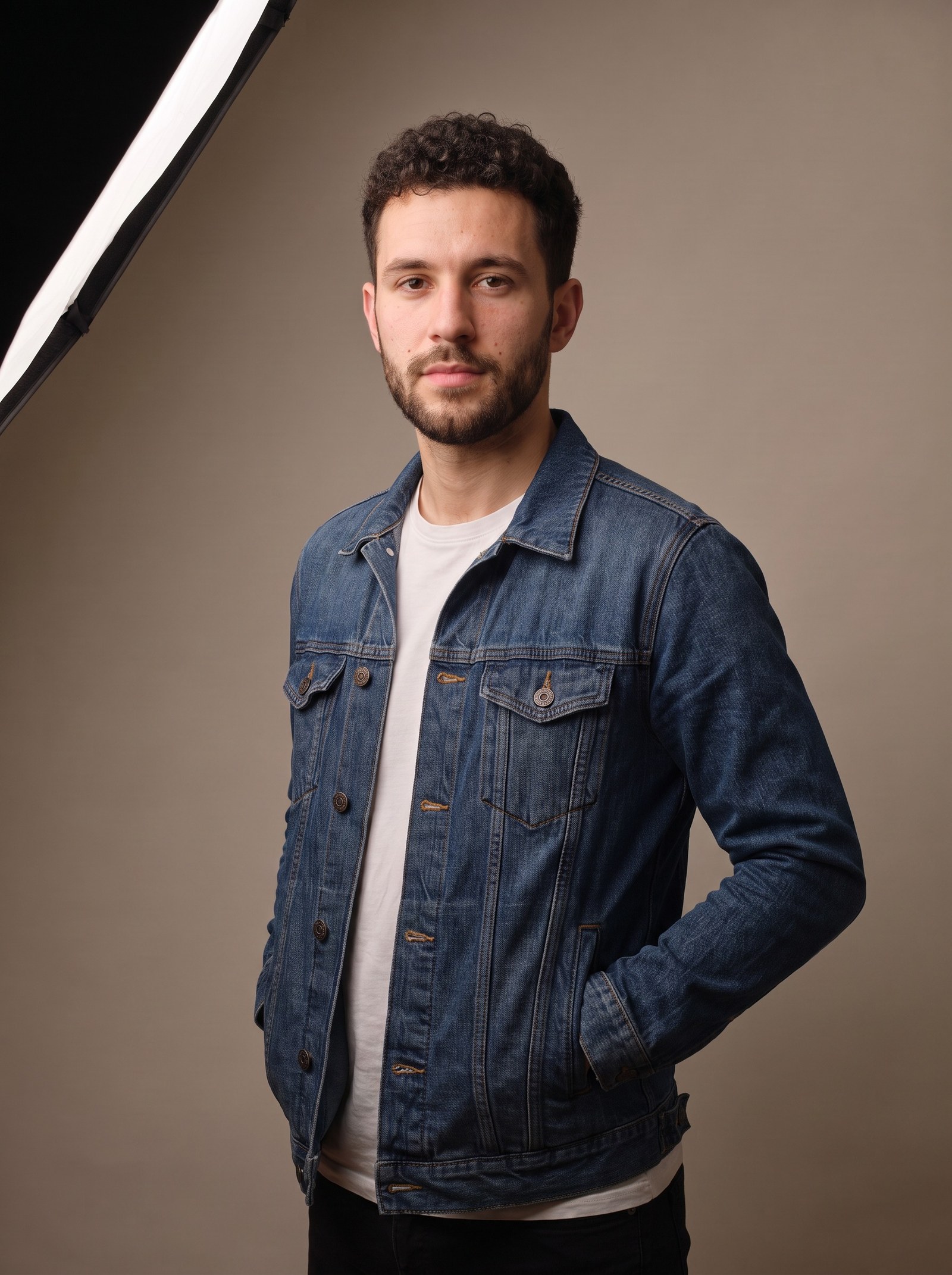 The same man with the same face, beard, denim jacket and backdrop, now turned into a three-quarter stance with his hands in his jacket pockets, under the same softbox in the same studio light