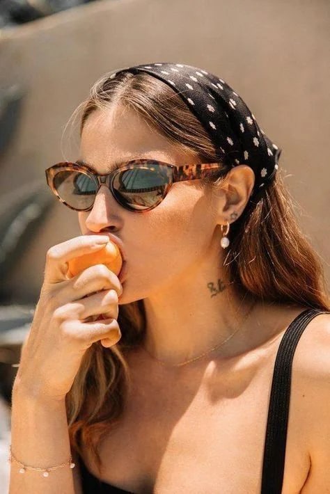 Small, soft phone snapshot of a woman in tortoiseshell sunglasses and a black polka-dot headscarf biting into a peach in bright sun — the original low-resolution file