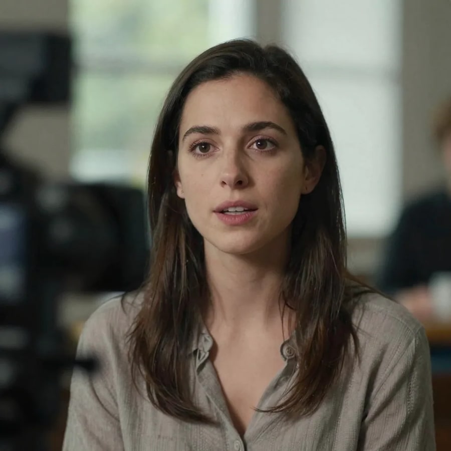 A woman speaking to camera in an interview setup, a camera body blurred in the foreground and a window behind her