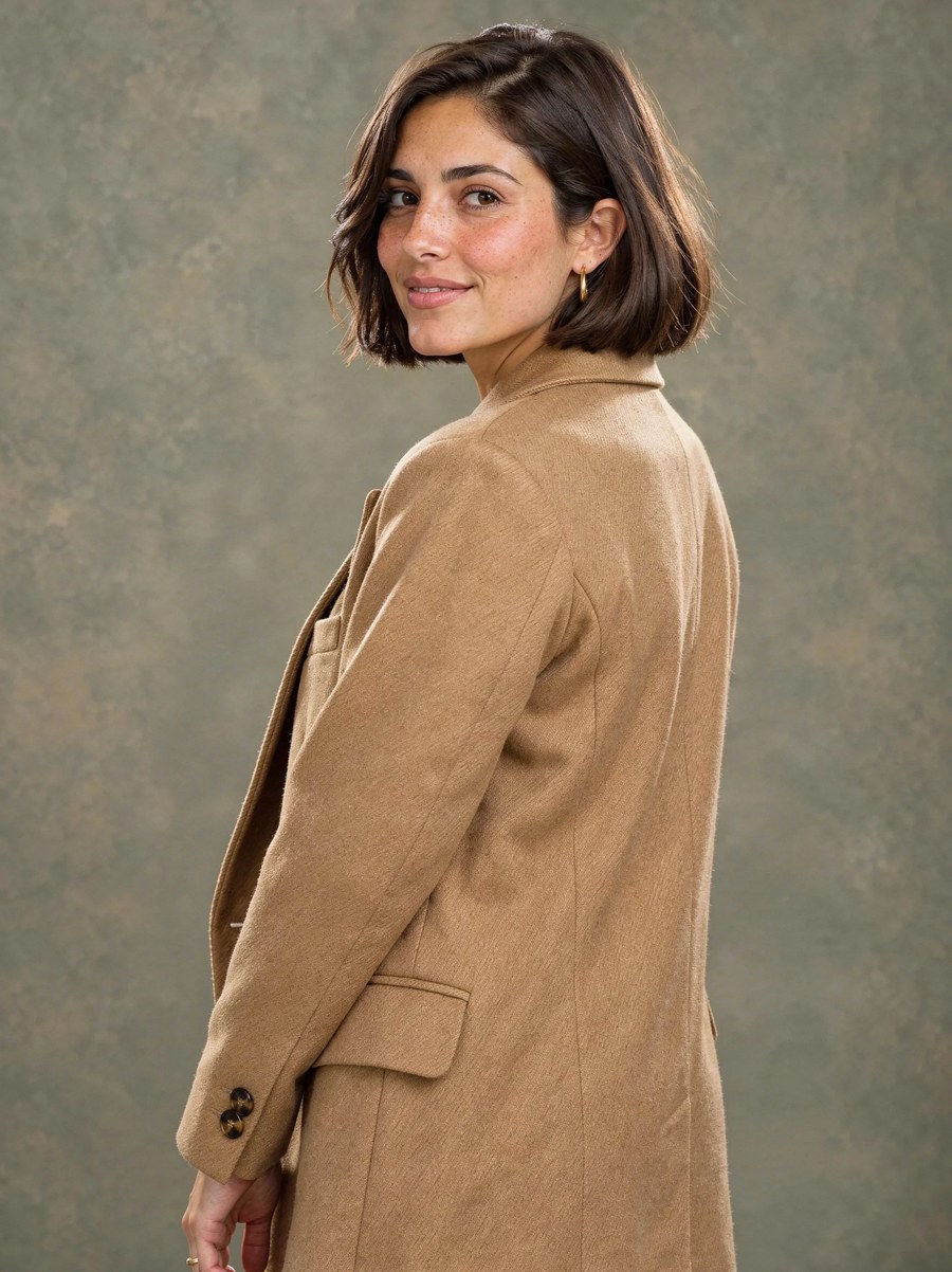 A woman with a chin-length dark brown bob and freckles, looking back over the shoulder of a camel wool coat, in front of a mottled olive studio backdrop