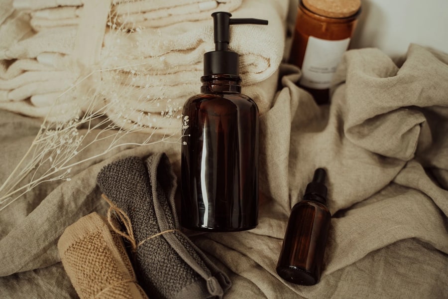 The product photo the reel is built from: an amber glass pump bottle with a black pump lying on rumpled linen beside a small amber dropper bottle, a cork-topped amber candle jar, folded cream towels, two rolled washcloths and dried gypsophila