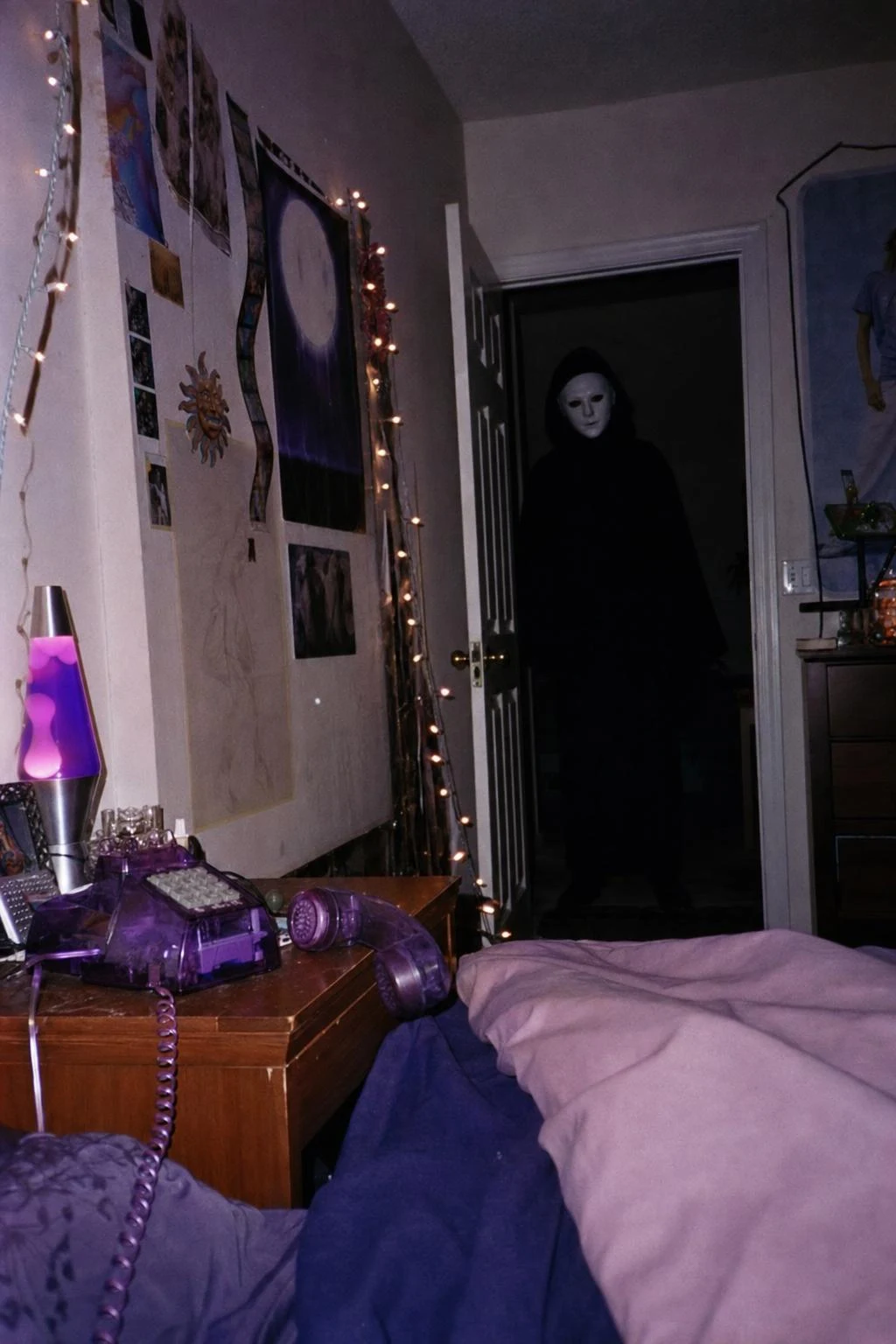 A flash photo of a Y2K-style bedroom at night with a purple landline off the hook and a figure in a plain white mask in the dark doorway